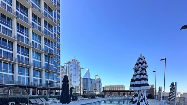 Área de piscina no terraço com espreguiçadeiras e um guarda-sol listrado, rodeada por edifícios modernos.