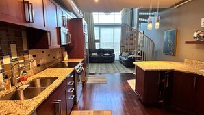 Cozinha moderna com bancadas de granito, eletrodomésticos de aço inoxidável e vista para uma sala de estar com sofá e escada.