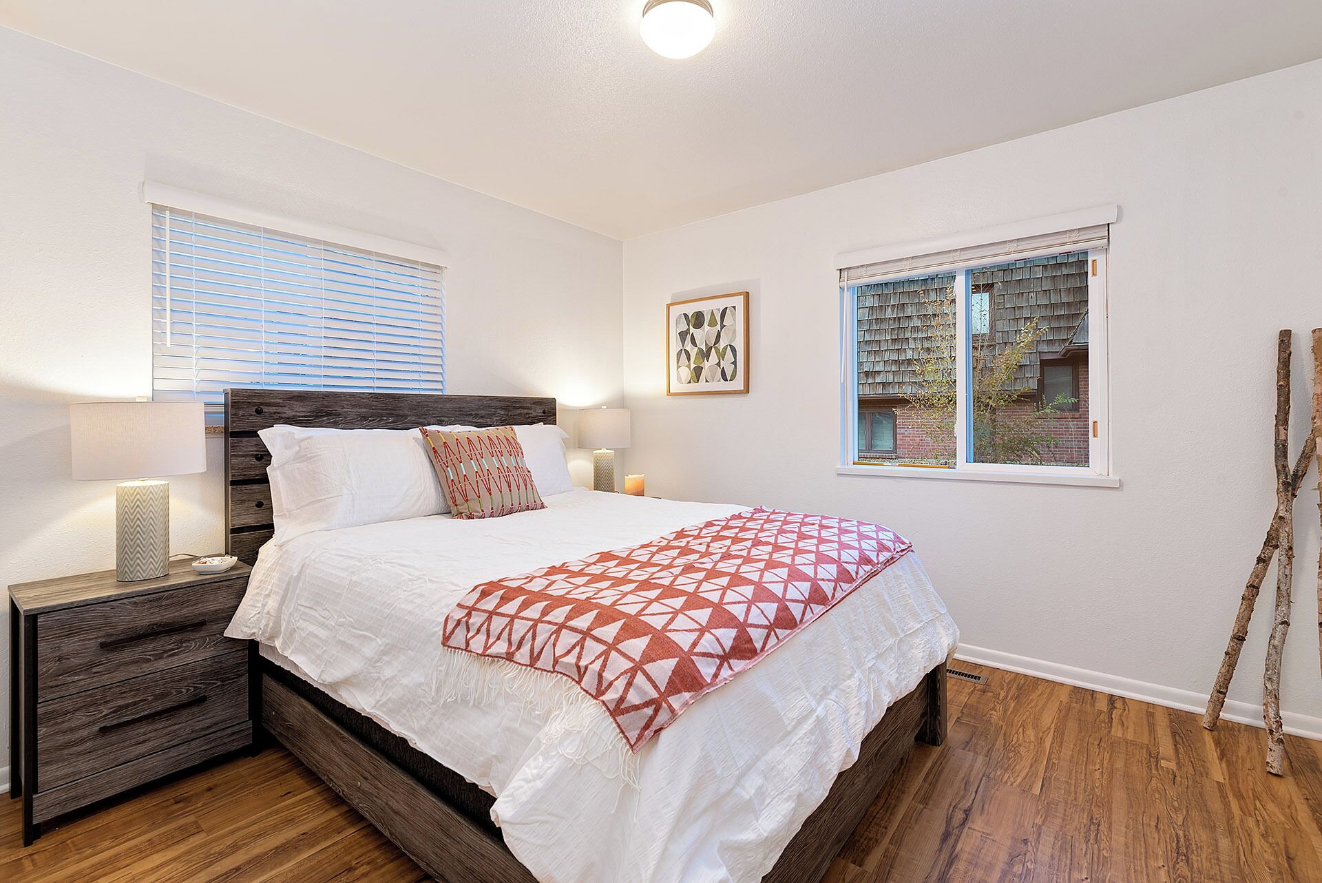 Une chambre à coucher avec un lit en bois, une literie blanche, une table de chevet avec une lampe et deux fenêtres équipées de stores.