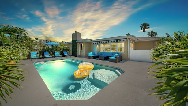A swimming pool with lounge chairs and umbrellas, surrounded by palm trees and a building with large windows. - Villa Paradisus. Heated Pool! 6 min to the beach (Satellite Beach)