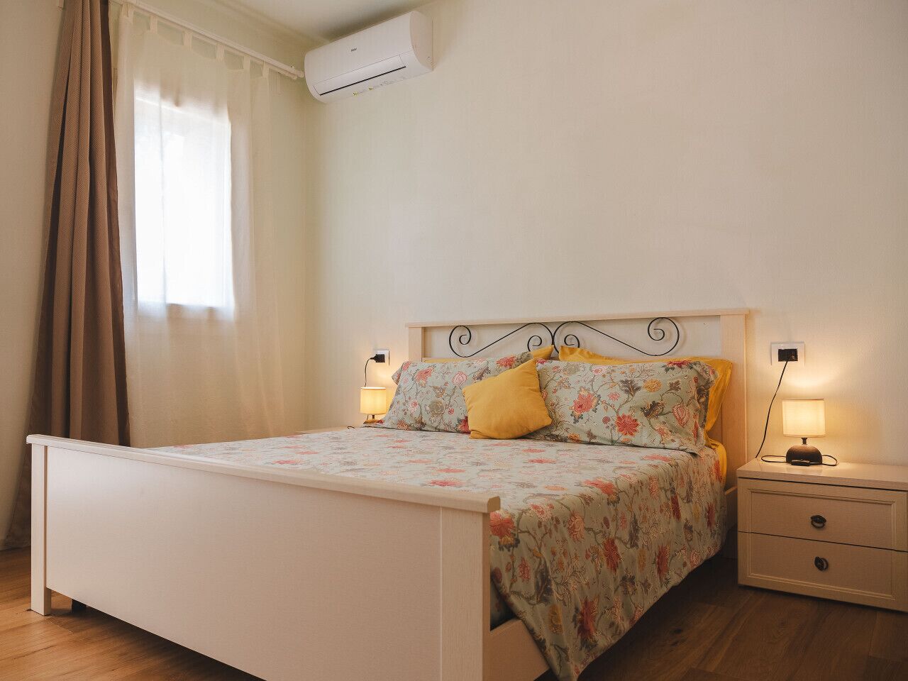 A bedroom with a bed, nightstands, a wall-mounted air conditioner, and a window with curtains.