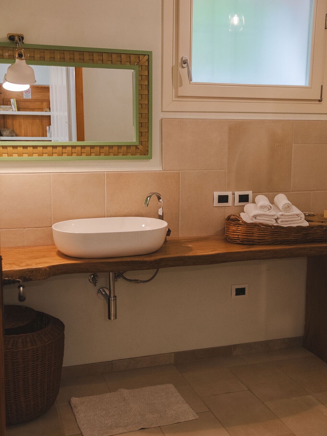 A bathroom with a white sink, a mirror, a window, and a basket of towels.