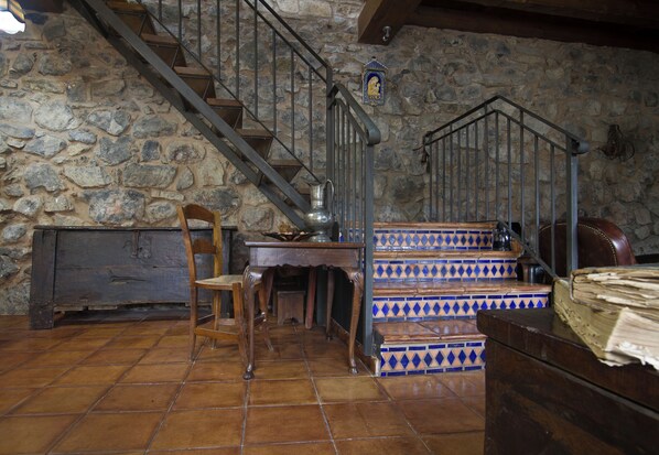 Una habitación rústica con pared de piedra, una mesa de madera, una silla, un banco y una escalera con azulejos azules y blancos.