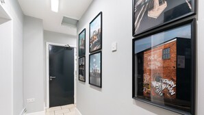 A hallway with framed pictures on the walls, a door, and a window with a brick wall and graffiti. - Citysteps Hotels Newcastle West (Newcastle-upon-Tyne)