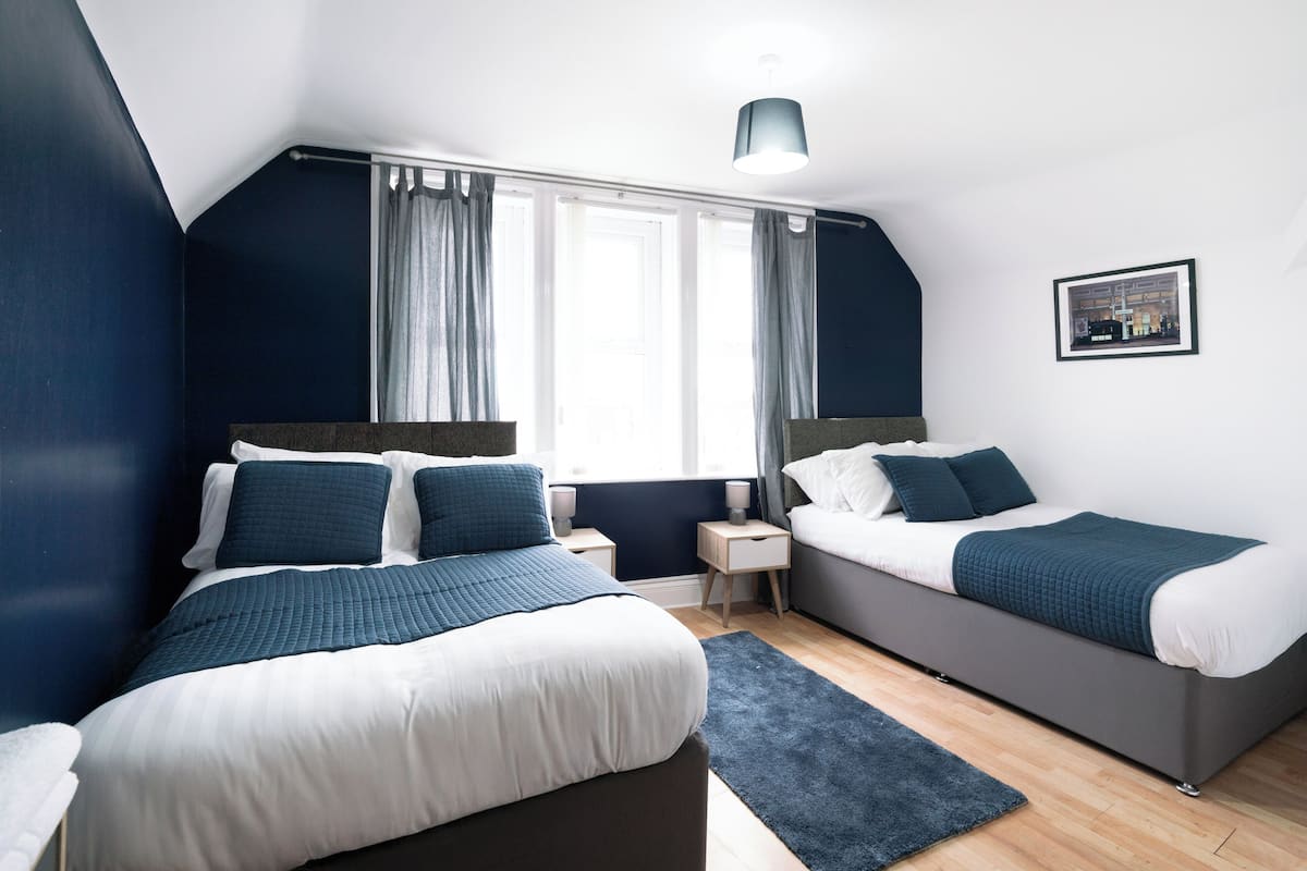 Une chambre moderne avec deux lits, une grande fenêtre avec des rideaux, une petite table de chevet et un luminaire au plafond.