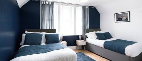 Un dormitorio moderno con dos camas, un ventanal con cortinas, una mesita de noche pequeña y una lámpara de techo.