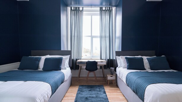 A bedroom with two beds, a desk, and a window with curtains. - Citysteps Hotels Newcastle West (Newcastle-upon-Tyne)