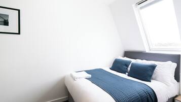 Una cama bien hecha con una manta y almohadas azules, una pared blanca y un cuadro enmarcado.
