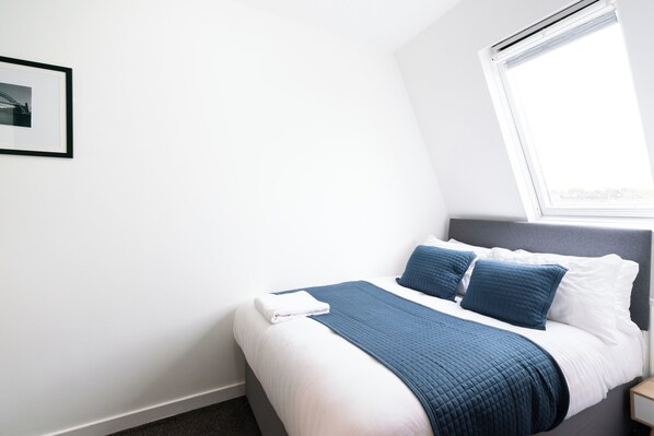Una cama bien hecha con una manta y almohadas azules, una pared blanca y un cuadro enmarcado.