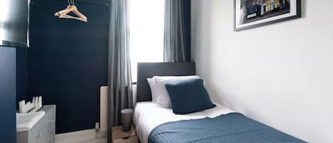 Une chambre à coucher comprenant un lit, une commode, un radiateur, une fenêtre avec des rideaux et un tableau encadré accroché au mur.