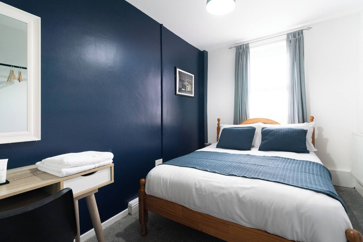 Une chambre à coucher comprenant un lit, une table de chevet, une chaise, une fenêtre avec des rideaux et un mur orné d’un tableau.