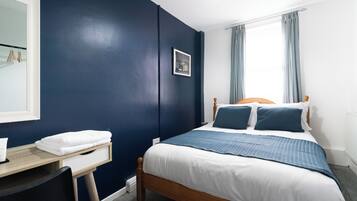 A bedroom with a bed, a bedside table, a chair, a window with curtains, and a wall with a picture.