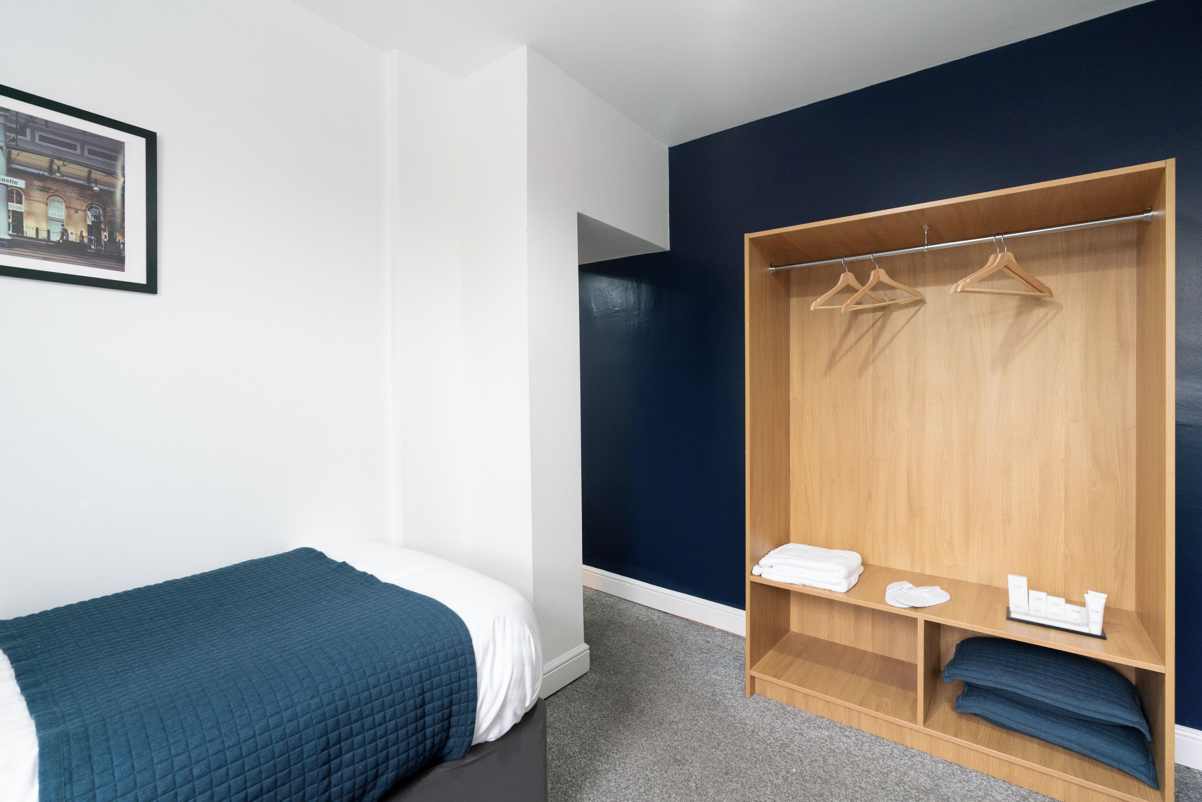 A bedroom with a single bed, a wooden wardrobe with hangers, a shelf with folded towels, and a framed picture on the wall.