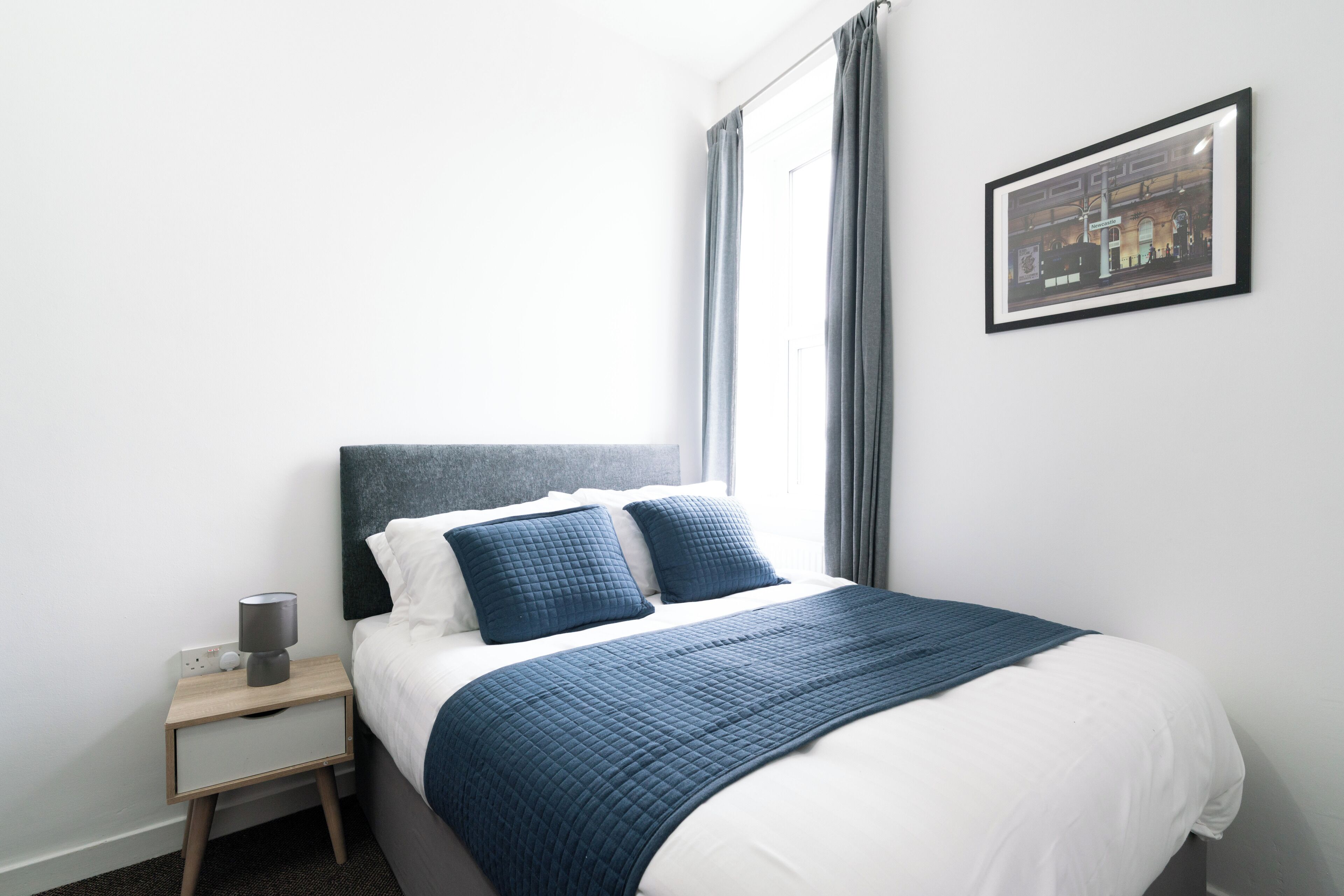 Una cama bien hecha con un edredón azul, una lámpara de noche y un cuadro en la pared.