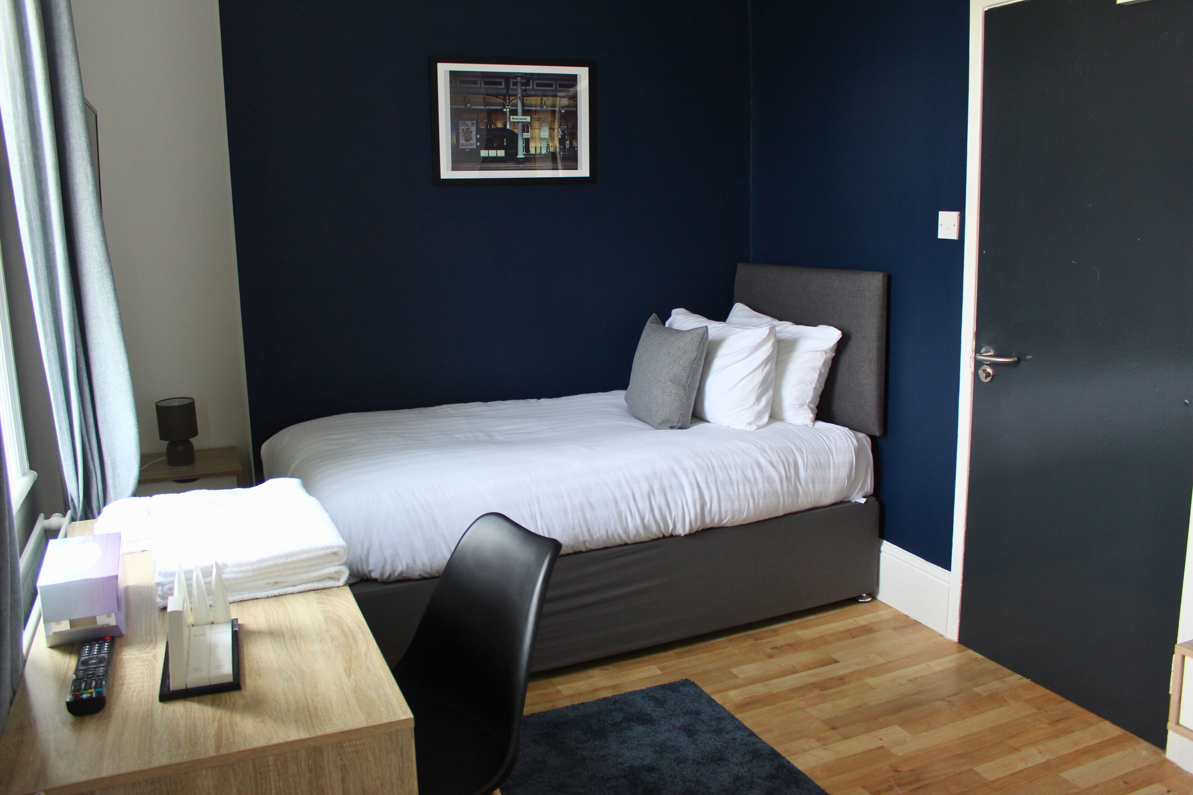 Una cama bien hecha con ropa de cama blanca y gris, una mesita de noche con un control remoto, una silla y un cuadro en la pared.