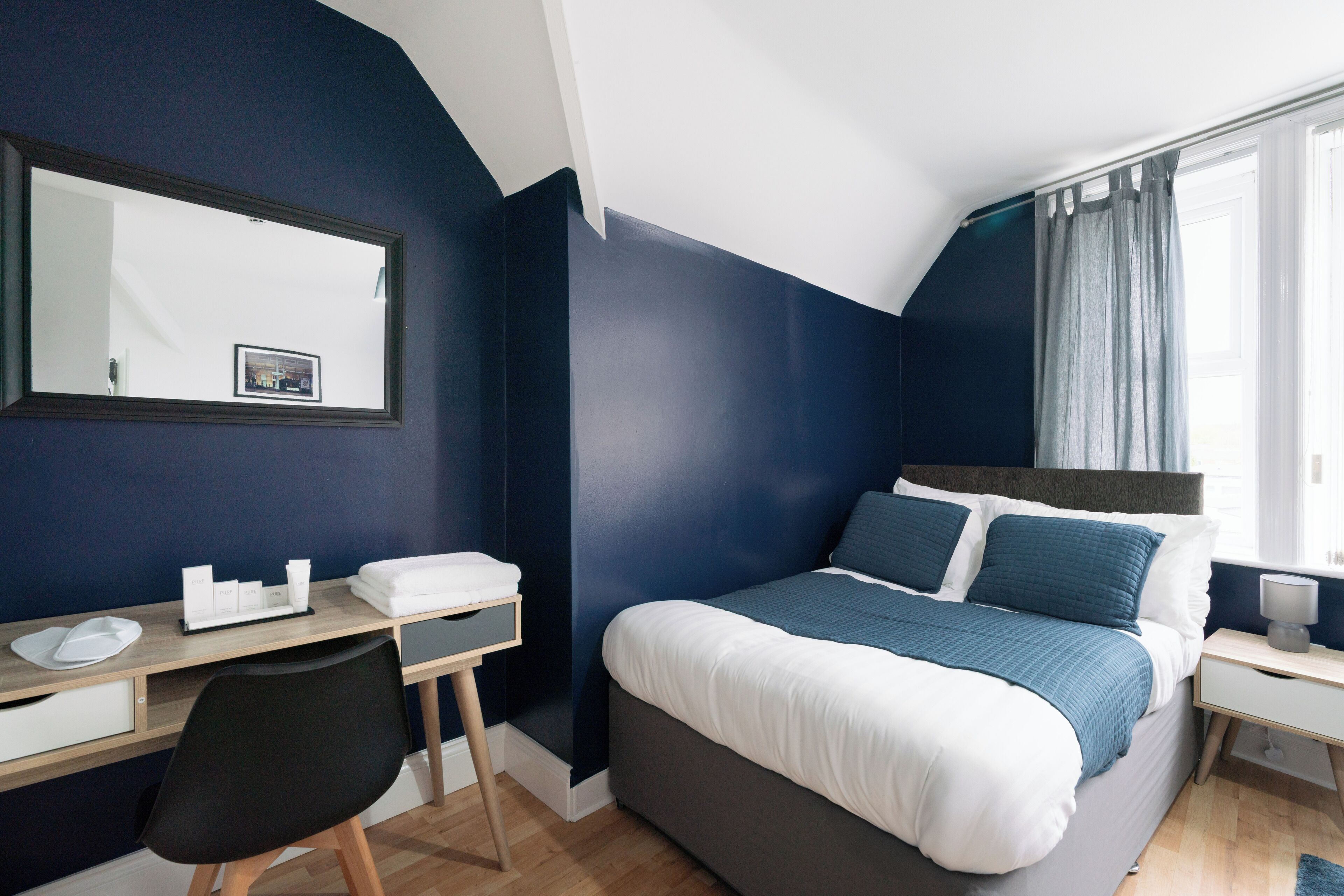 Une chambre à coucher comprenant un lit, un bureau, une chaise, une table de chevet et une fenêtre avec des rideaux.
