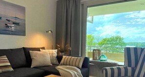 Un salon moderne avec un canapé, des coussins, une lampe sur pied et une vue sur un balcon doté d’une balustrade et de plantes vertes.
