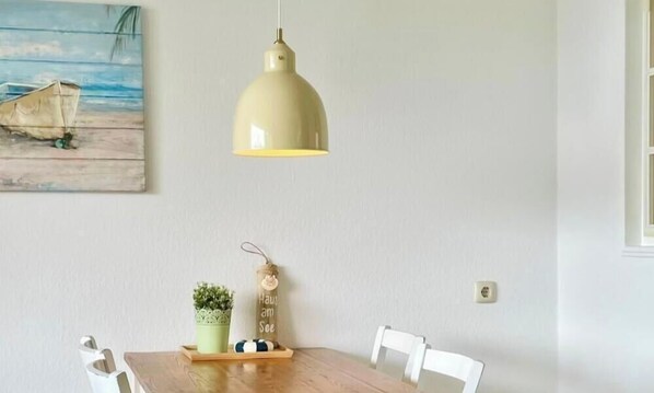 Un coin repas avec une table en bois, des chaises blanches, une suspension, une petite plante en pot et un objet décoratif posé sur la table.