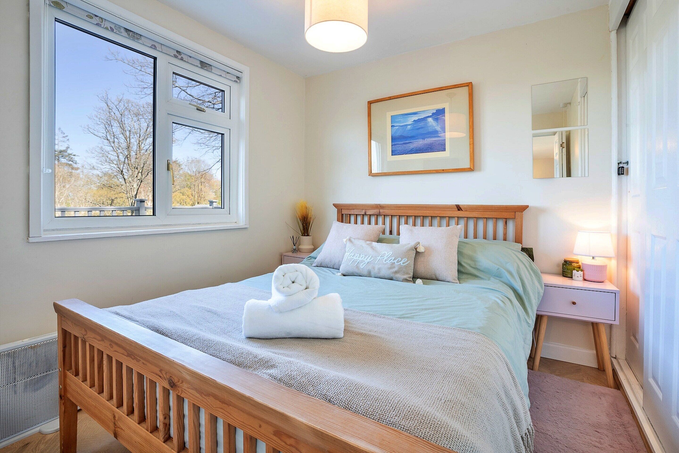 Un dormitorio con cama de madera, una mesita de noche, una lámpara, un cuadro enmarcado en la pared y una ventana con vistas a árboles.