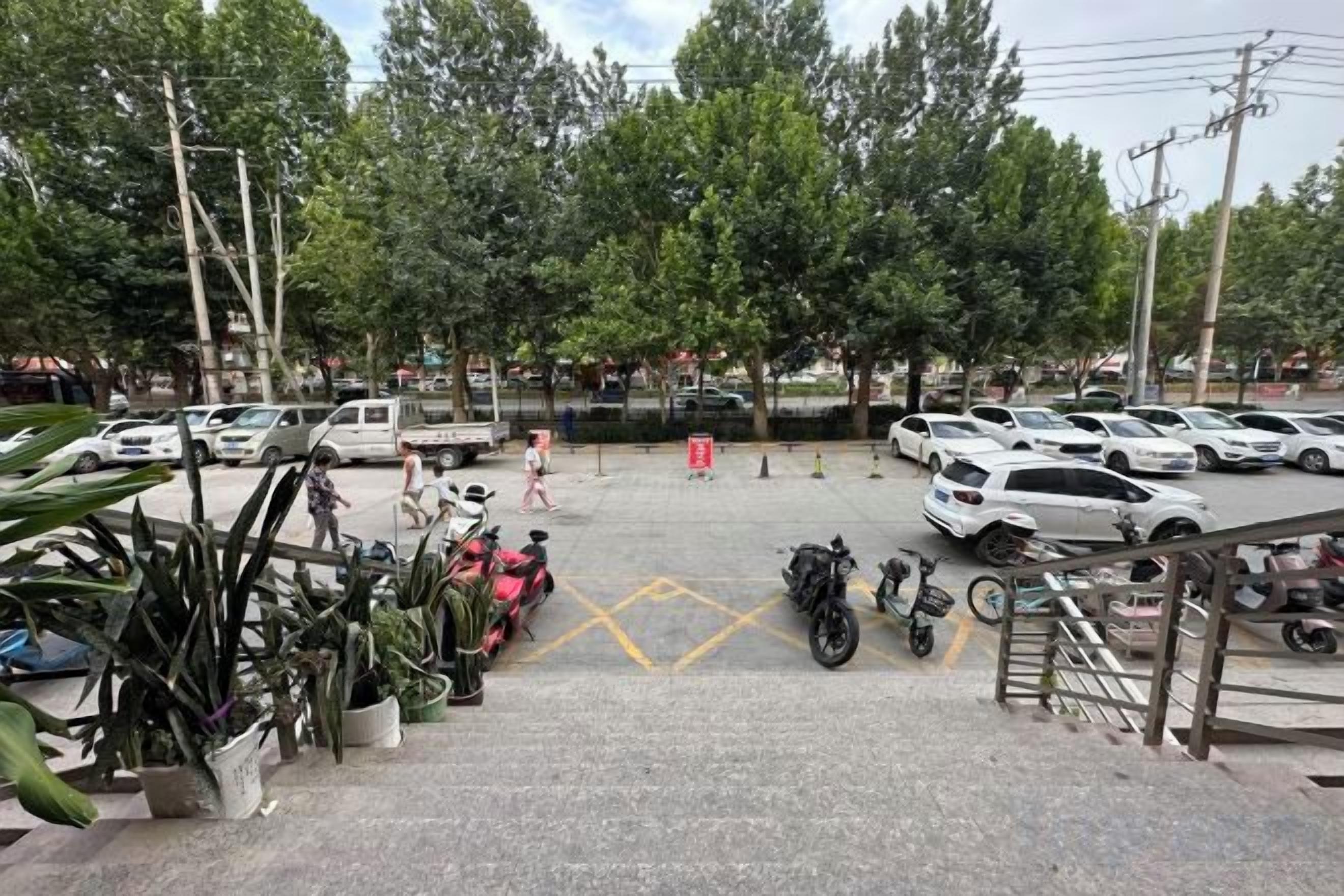 A parking lot with multiple cars and motorcycles, a staircase leading to a building, and potted plants.