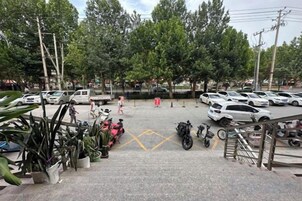 Un estacionamiento con varios coches y motocicletas, una escalera que conduce a un edificio y plantas en macetas.