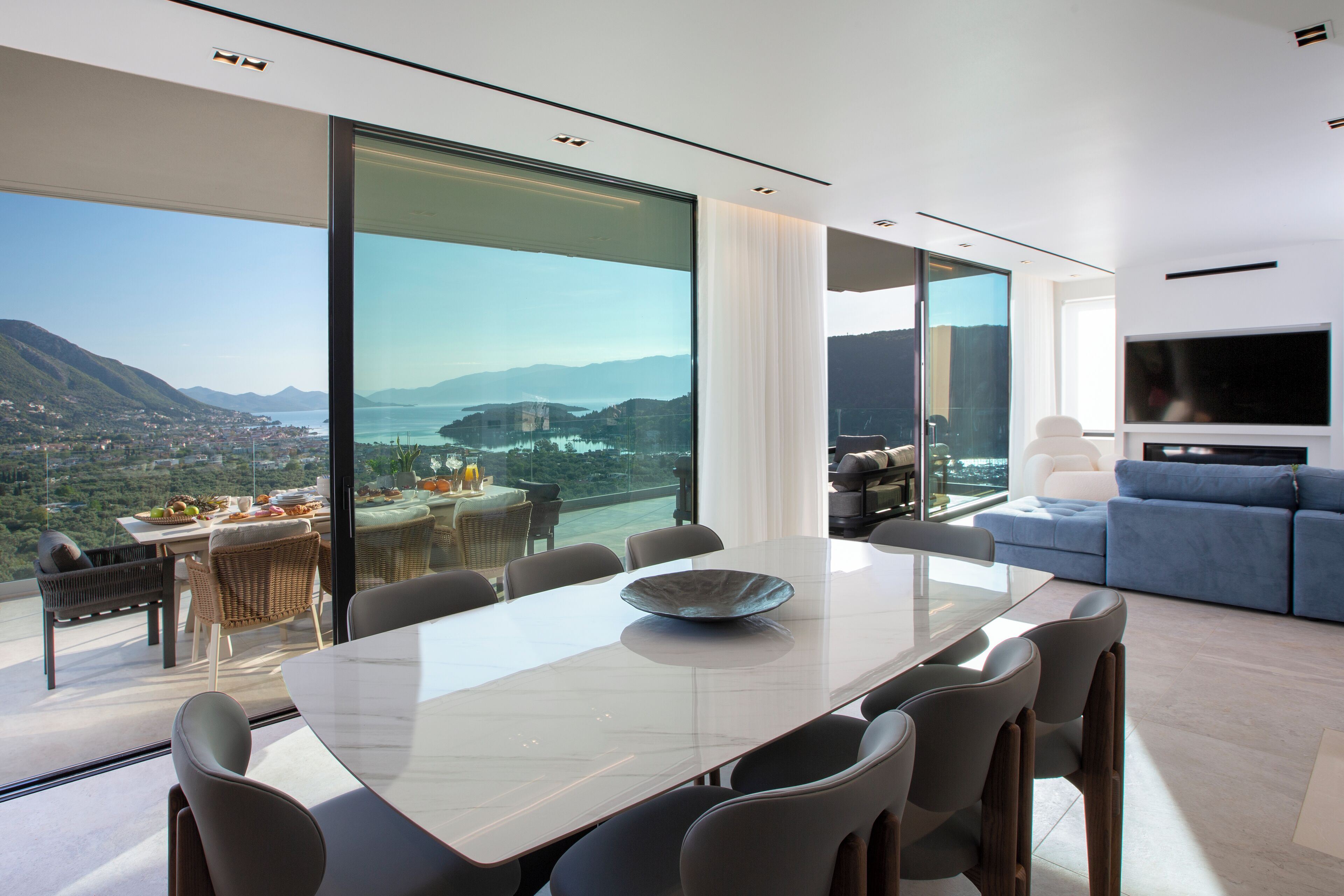 Un comedor moderno con una mesa grande y sillas, un cuenco, y una vista panorámica de un lago y montañas a través de amplias ventanas.