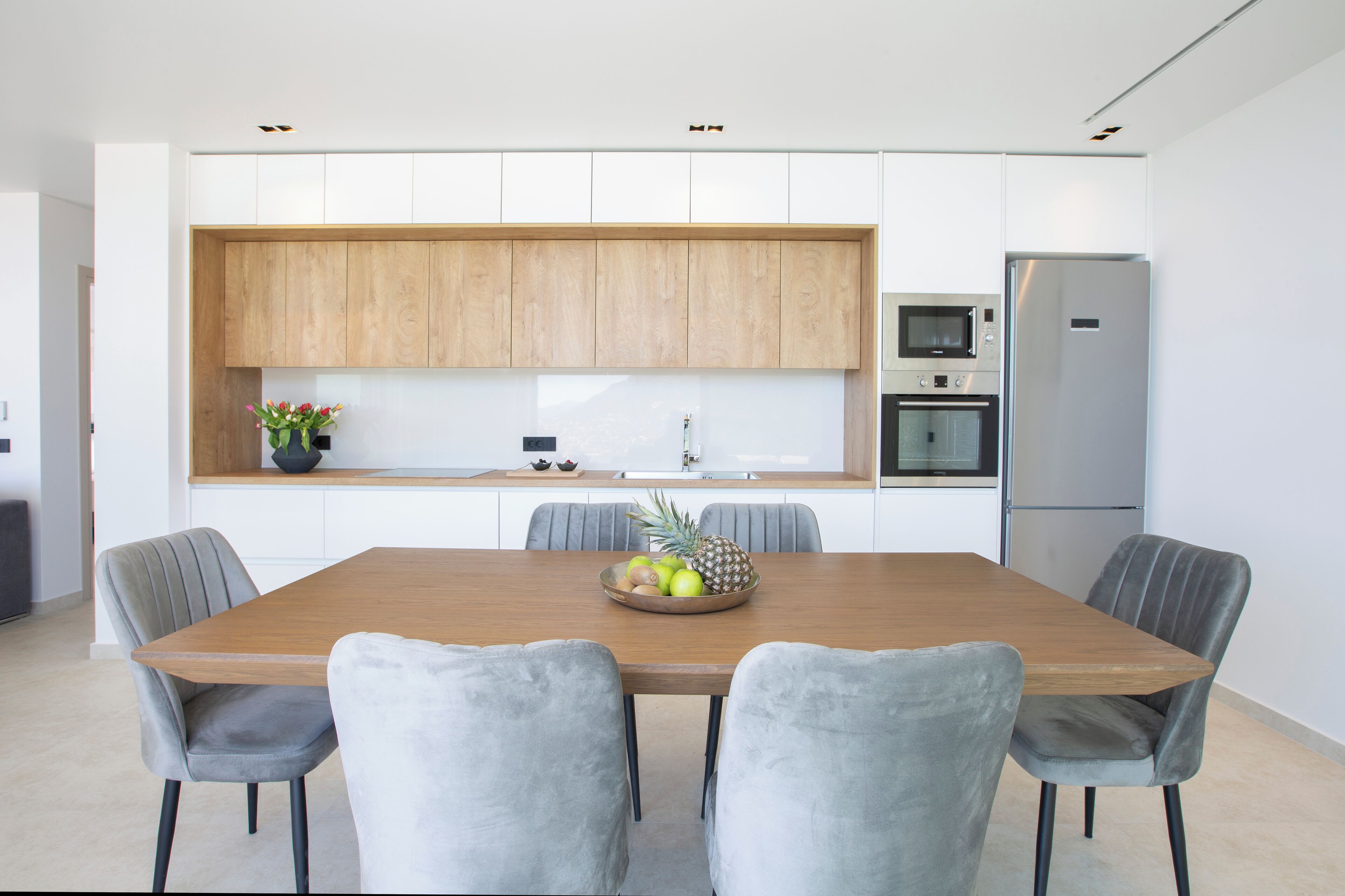A modern kitchen with a wooden dining table, grey upholstered chairs, and a fruit bowl.