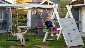 Zwei Kinder schaukeln in einem Garten mit einer Kletterwand und einem Grillbereich.