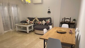 Un salon moderne avec une table à manger en bois, des chaises blanches, un canapé gris agrémenté de coussins à motifs floraux, une petite table basse blanche et une décoration murale.