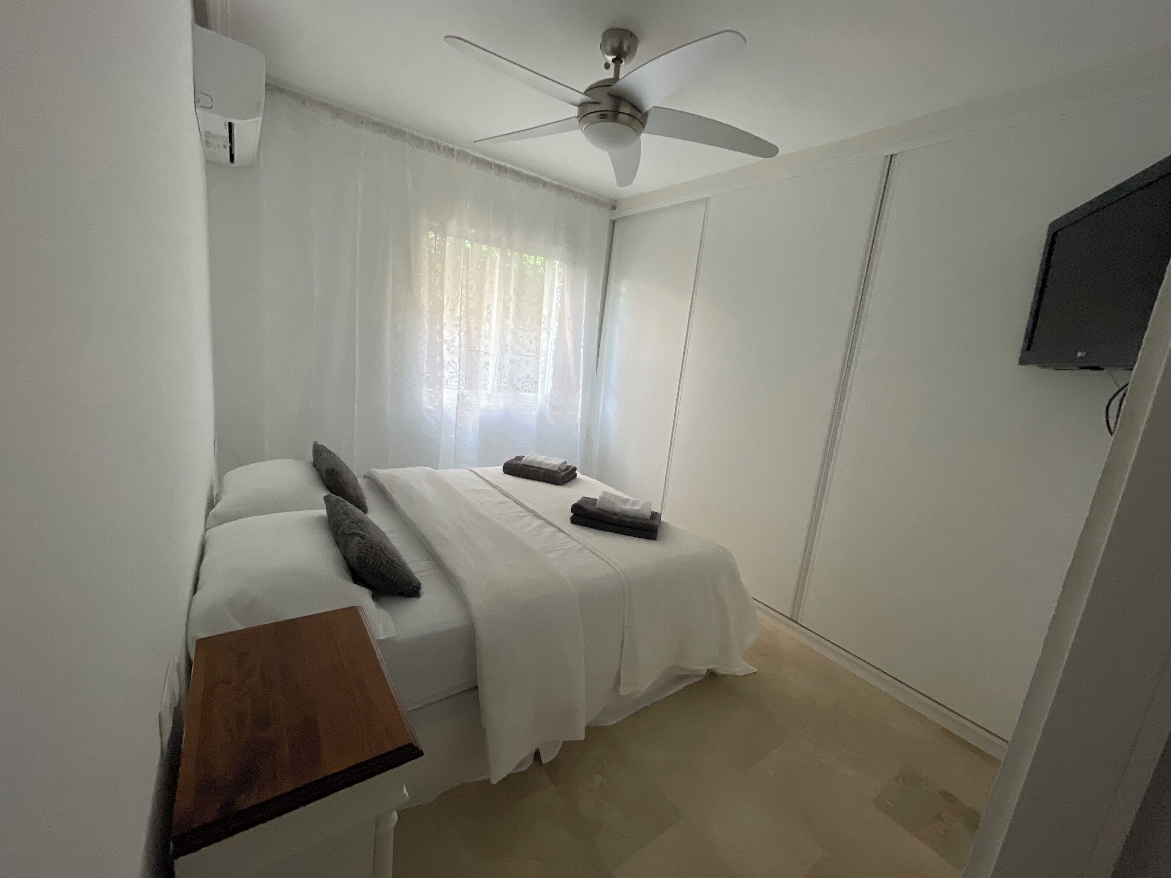 A bedroom with a bed, a ceiling fan, a TV mounted on the wall, and a window with sheer curtains.