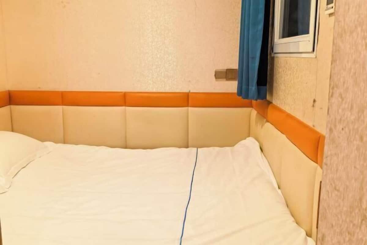 A bed with a white mattress and orange headboard, a window with a blue curtain, and a wall-mounted light switch.