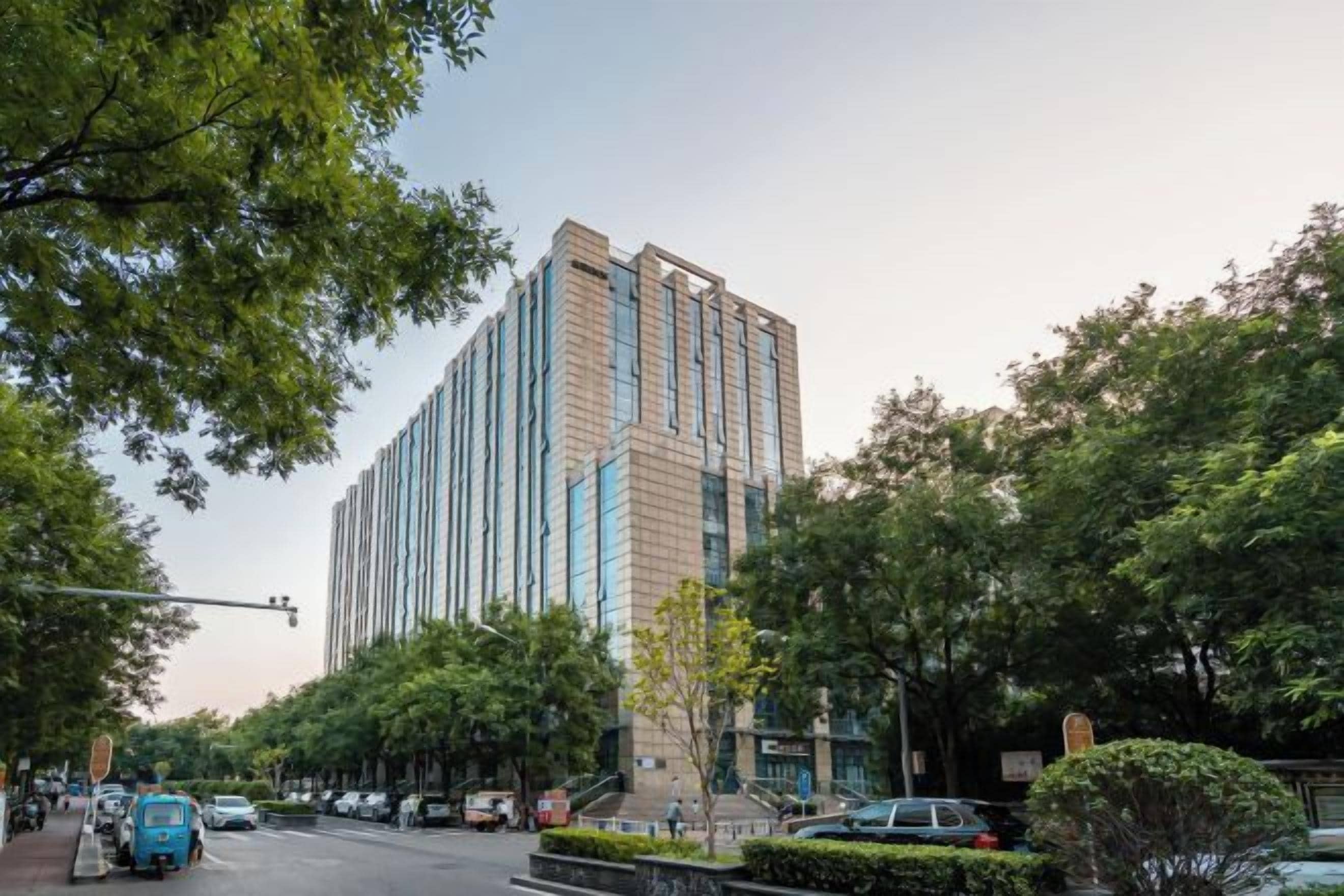 A tall building with a modern design, surrounded by trees and parked cars on a street.