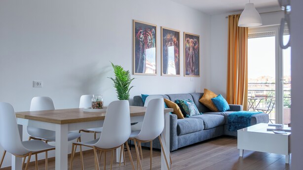 A modern living room with a dining area, featuring a grey sofa, white chairs, and a wooden dining table. There are two framed artworks on the wall and a potted plant. - Between sea and city, 15 minutes from the airport, 5 minutes from the excavations of Ostia Antina, (Lazio)