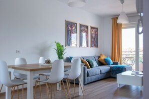 A modern living room with a dining area, featuring a grey sofa, white chairs, and a wooden dining table. There are two framed artworks on the wall and a potted plant. - Between the sea and the city, 15 minutes from the airport, 5 minutes from the archaeological site of Ostia Antica, (Lazio)