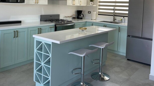 Een moderne keuken met lichtblauwe kastjes, een wit kookeiland met barkrukken, roestvrijstalen apparatuur en een raam met zonwering.