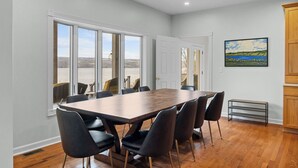 A dining area with a large wooden table and black chairs, large windows with a view of a landscape, and a painting on the wall. - Lakefront 6BR Retreat with Private Dock, Huge Deck & Epic Seneca Views (Dundee)