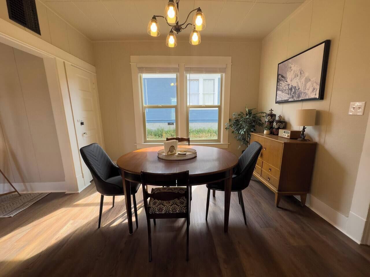 A dining area with a round table, four chairs, a chandelier, a cabinet, and a window with a view.