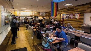 A busy cafeteria with people dining at tables, a counter with food items, and a rainbow flag hanging from the ceiling. - Downtown Studio | Small + Stylish | Near WWU (Bellingham)