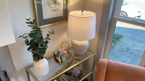 A room with a glass and gold side table, a white vase with green leaves, a basket with books, a lamp, and a pink chair. - Downtown Studio | Small + Stylish | Near WWU (Bellingham)