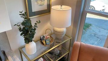 A room with a glass and gold side table, a white vase with green leaves, a basket with books, a lamp, and a pink chair.