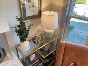 A room with a glass and gold side table, a white vase with green leaves, a basket with books, a lamp, and a pink chair.