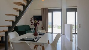 Un espace repas moderne avec une table blanche dressée pour quatre personnes, un escalier avec des marches en bois, un canapé vert et une vue sur l’extérieur grâce à de grandes fenêtres.