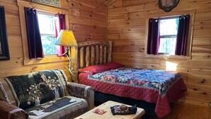 A cozy cabin room with a wooden interior, a bed with a quilt, a sofa with a floral blanket, and a table with a notebook and pen. - Pine Ridge Cabin - A Cozy Getaway for Two near Jasper AR! (Jasper)