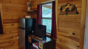 A compact wooden cabin with a kitchenette, a flat-screen TV on a wooden stand, and a window with curtains. - Pine Ridge Cabin - A Cozy Getaway for Two near Jasper AR! (Jasper)