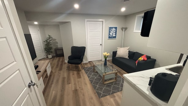 A compact living space with a wooden floor, a black armchair, a sofa, a glass coffee table, and a kitchen area with a countertop and sink. - The Snug Spot,Retreat offers a warm and inviting escape for travelers. (Winnipeg)