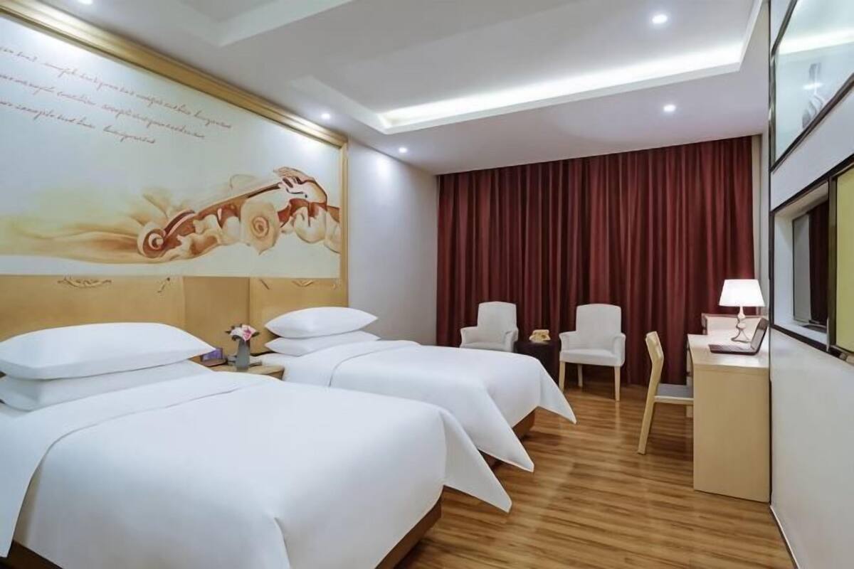 A hotel room with two beds, a desk with a lamp, a chair, and a large floral mural on the wall.