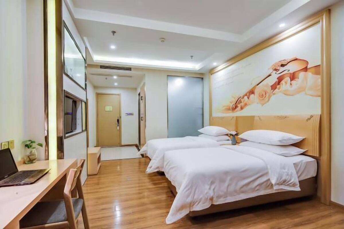 A hotel room with two beds, a desk with a laptop, a painting on the wall, and a hallway leading to another room.