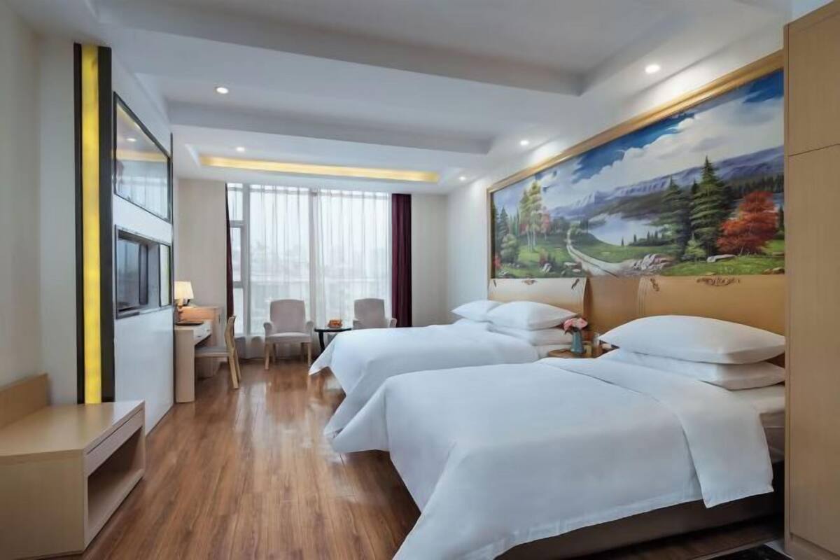 A hotel room with two beds, a desk, a chair, a TV, and a large landscape painting on the wall.