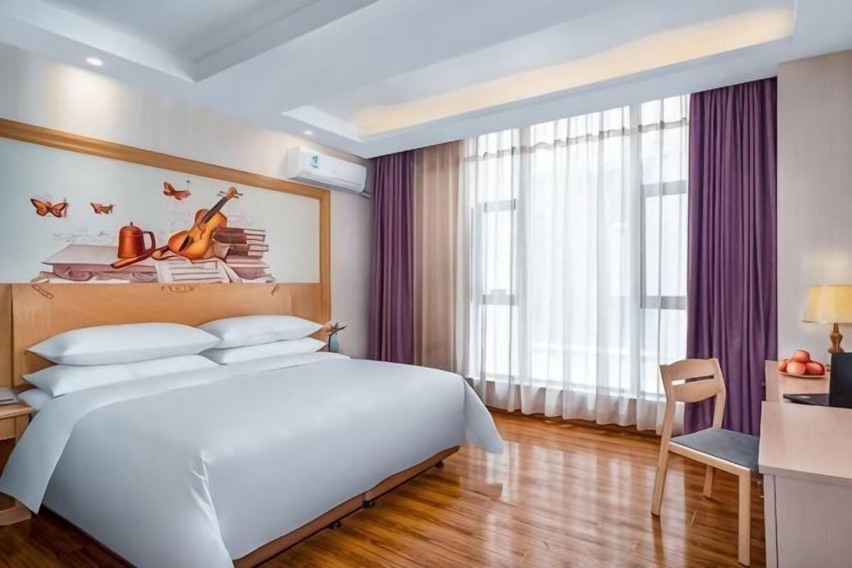 A hotel room with a large bed, a desk with a chair, a window with curtains, and a wall with a mural.