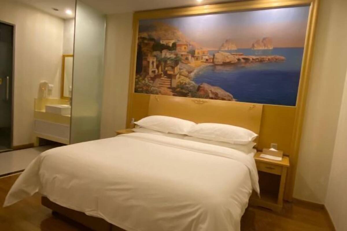 A hotel room with a bed, a nightstand, a painting of a coastal scene, and a bathroom with a glass shower enclosure.